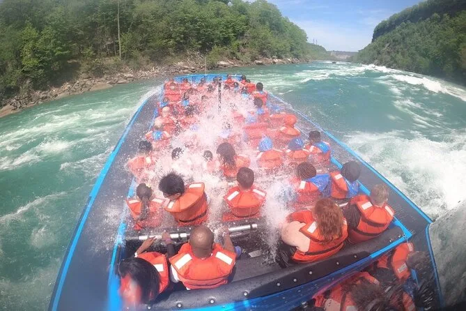 Mitfahrende in orangefarbenen Schwimmwesten in einem offenen Jetboot, das auf einem grünen Fluss in eine Welle prescht